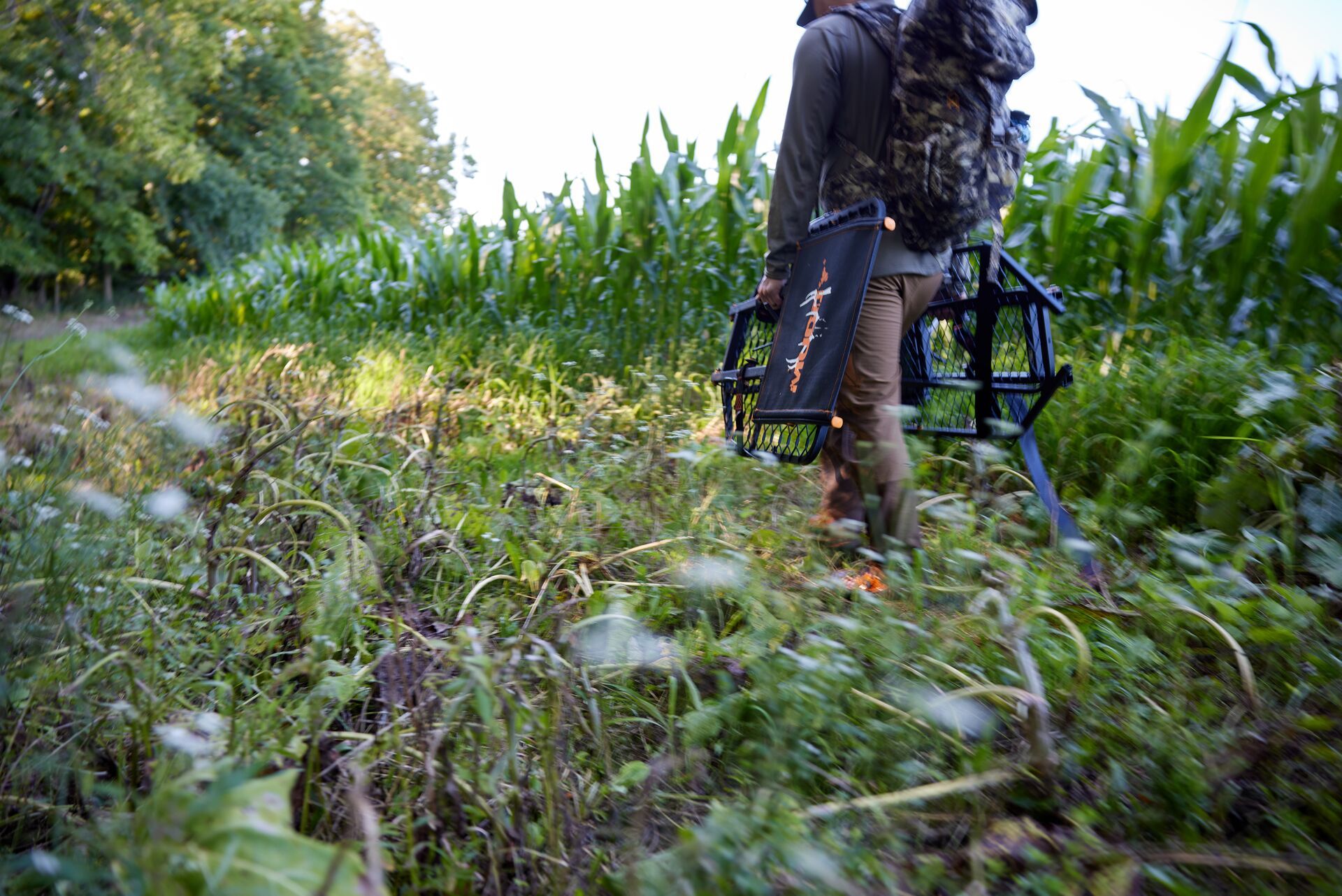 A hunter carrying gear and a tree stand in the brush near trees, avoid ticks pre-season prep. 