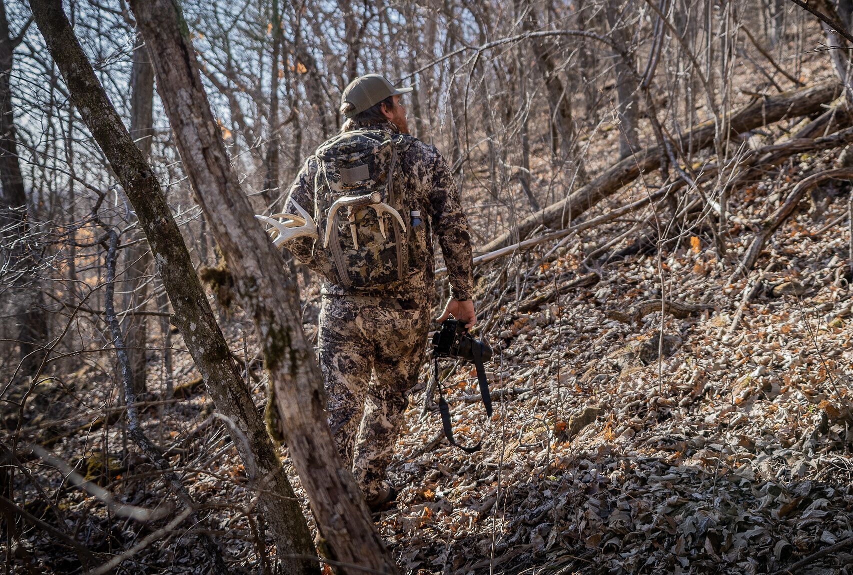 A hunter in camo walks in the woods with a camera, scouting for fall hunting season concept. 