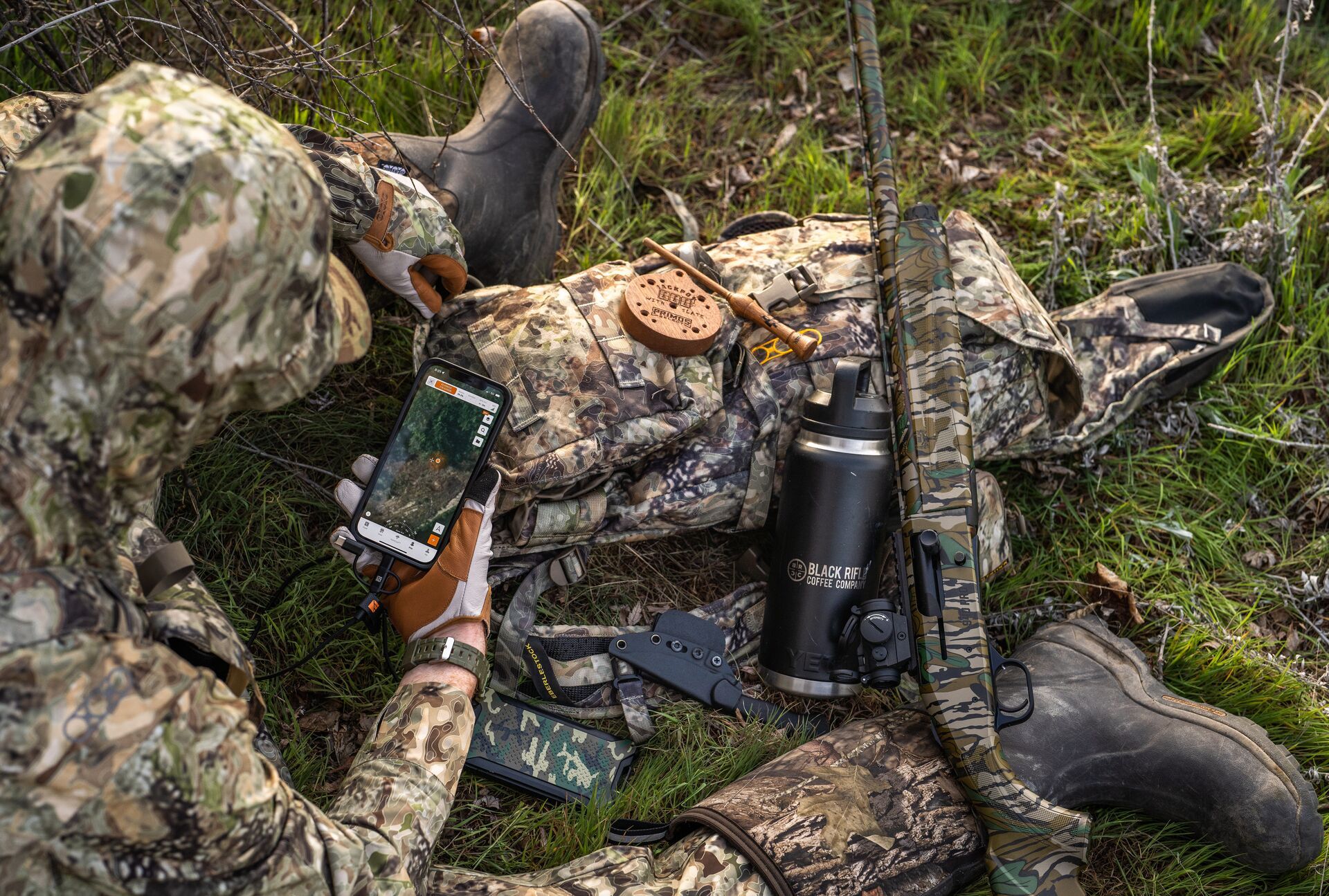 A hunter sits on the ground with a rifle, gear, and a phone showing the HuntWise app, Missouri hunting concept. 