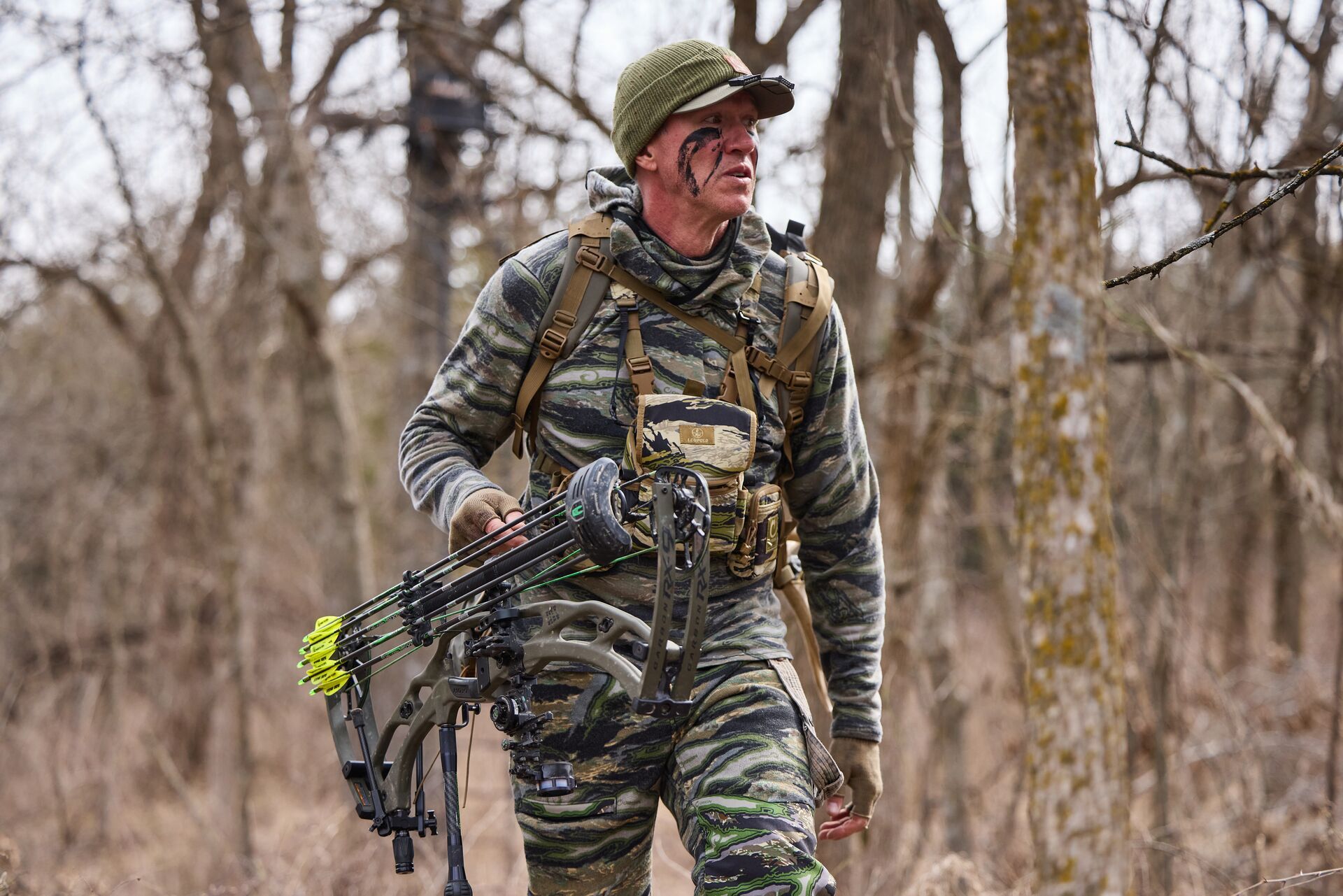 John Dudley in camo walks in woods with bow, how to get better at archery concept.