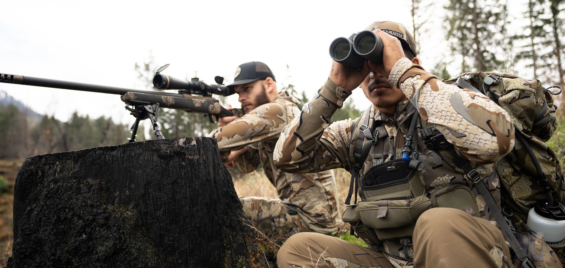 Hunter aims a rifle next to a hunter with binoculars, hunting shooting positions concept. 