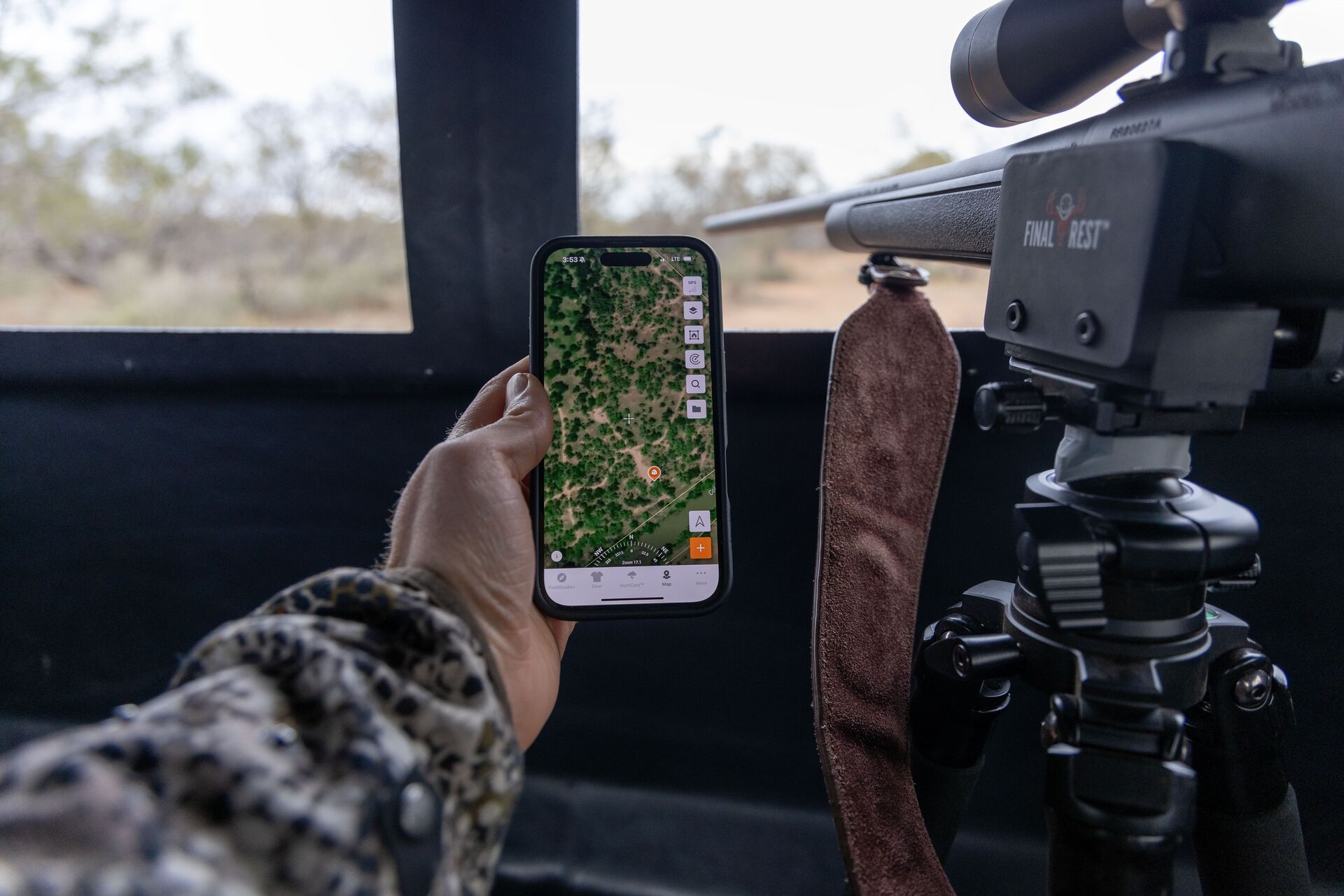 Close-up of a hand holding a phone showing the HuntWise app, with a rifle in a blind. 