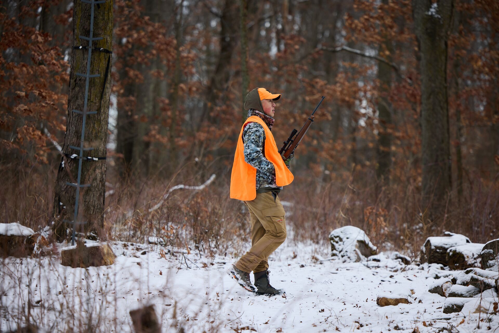 A hunter walks through snowy woods wearing camo and blaze orange while carrying a rifle.  