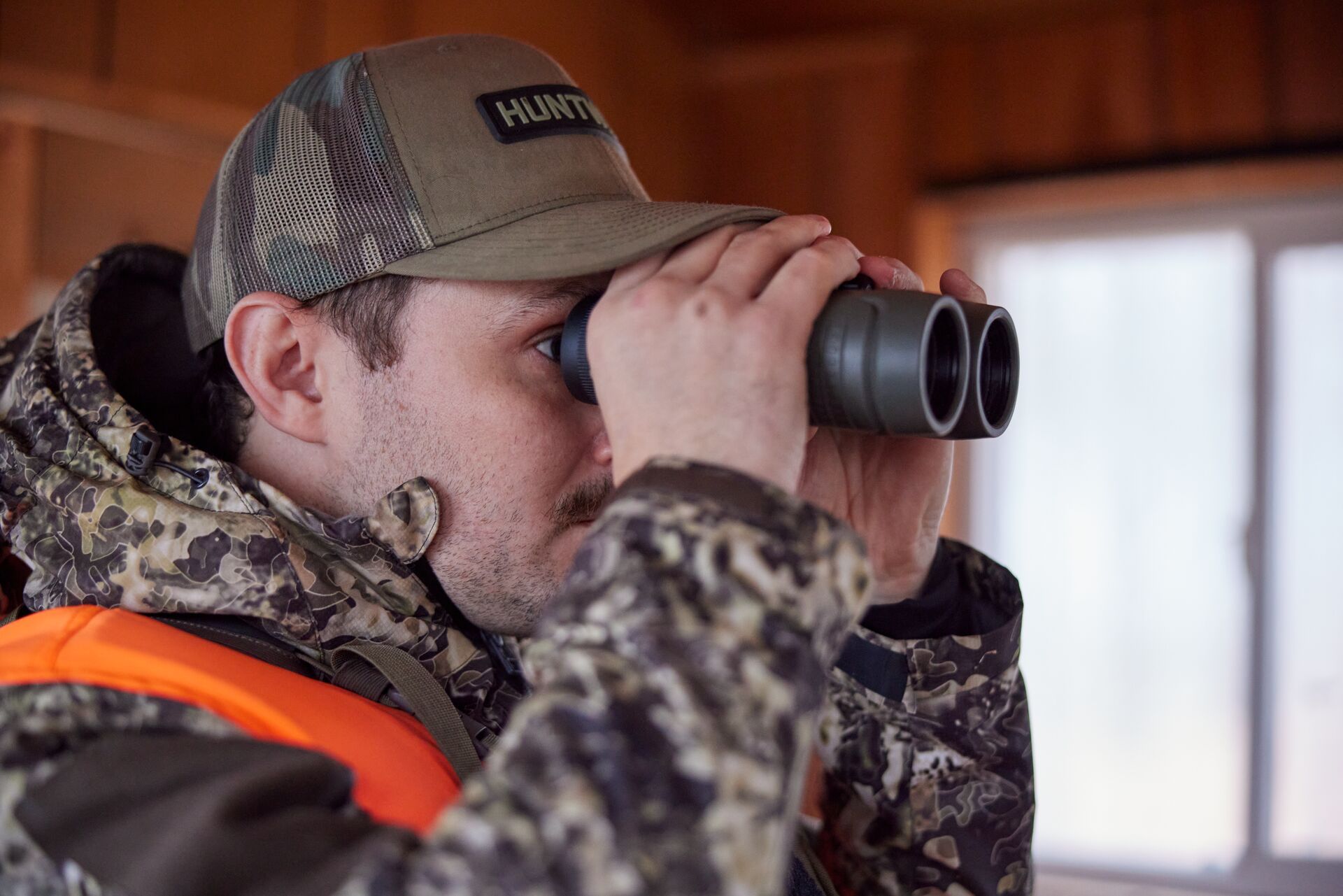 A hunter in camo and cold-weather gear looking through binoculars, deer hunting in January concept. 