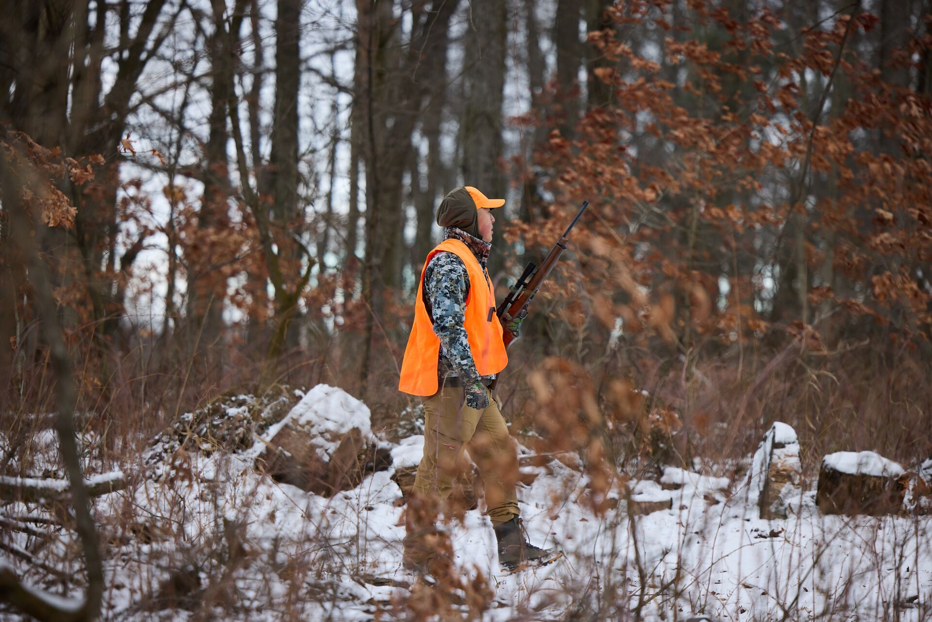 Hunter in blaze orange with rifle walks through snowy woods, beginner’s guide to small game hunting.