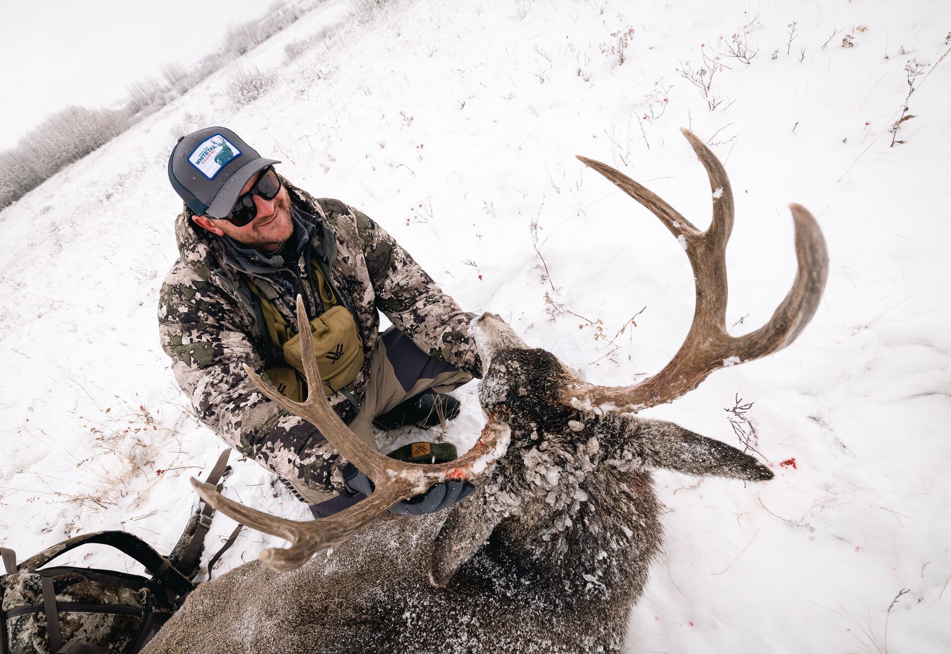 Hunter sits with buck deer in snow after hunt. 