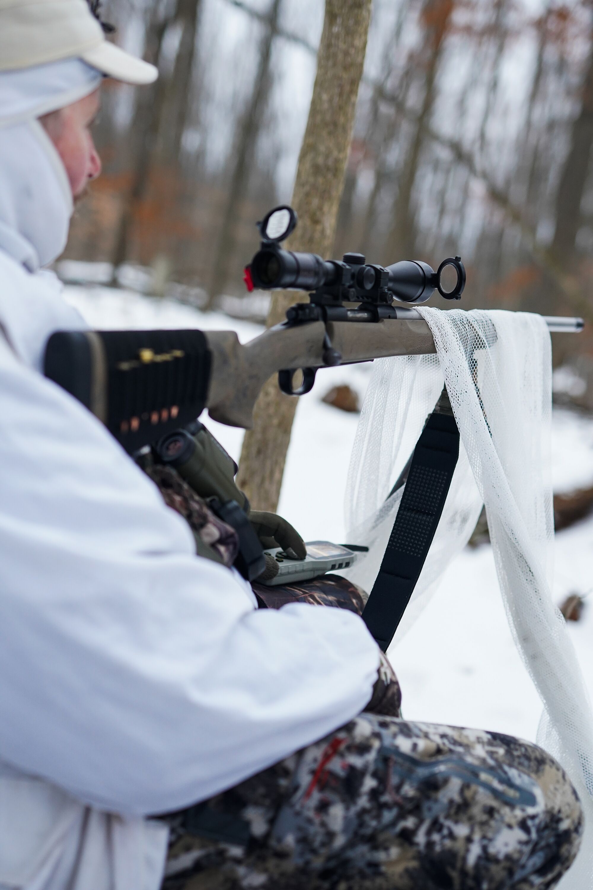 Hunter in white with rifle set up for hunting coyote in the winter.