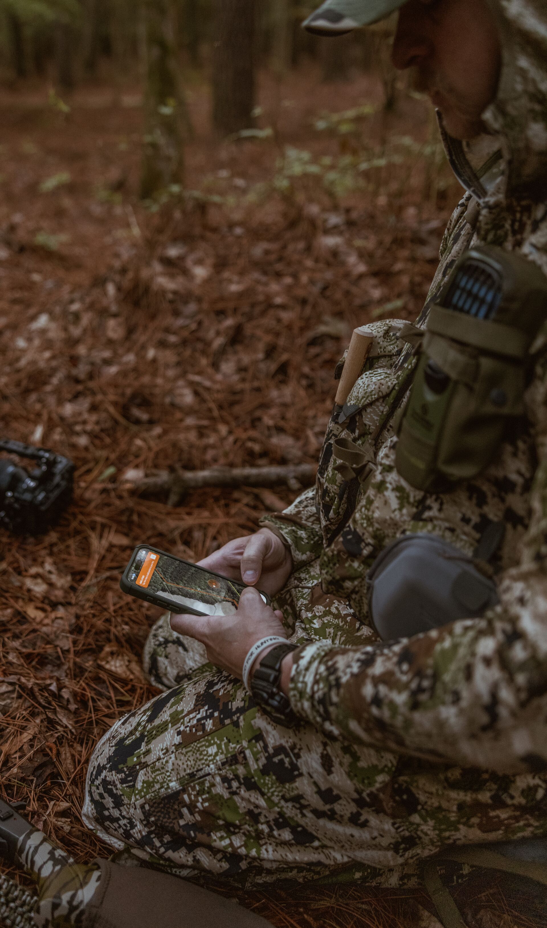 Hunter kneels in the woods using HuntWise on phone. 