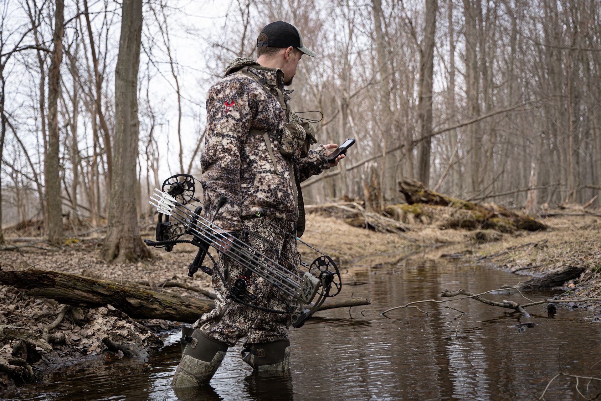 Hunter with bow walks through creek using HuntWise turkey hunting app on phone. 