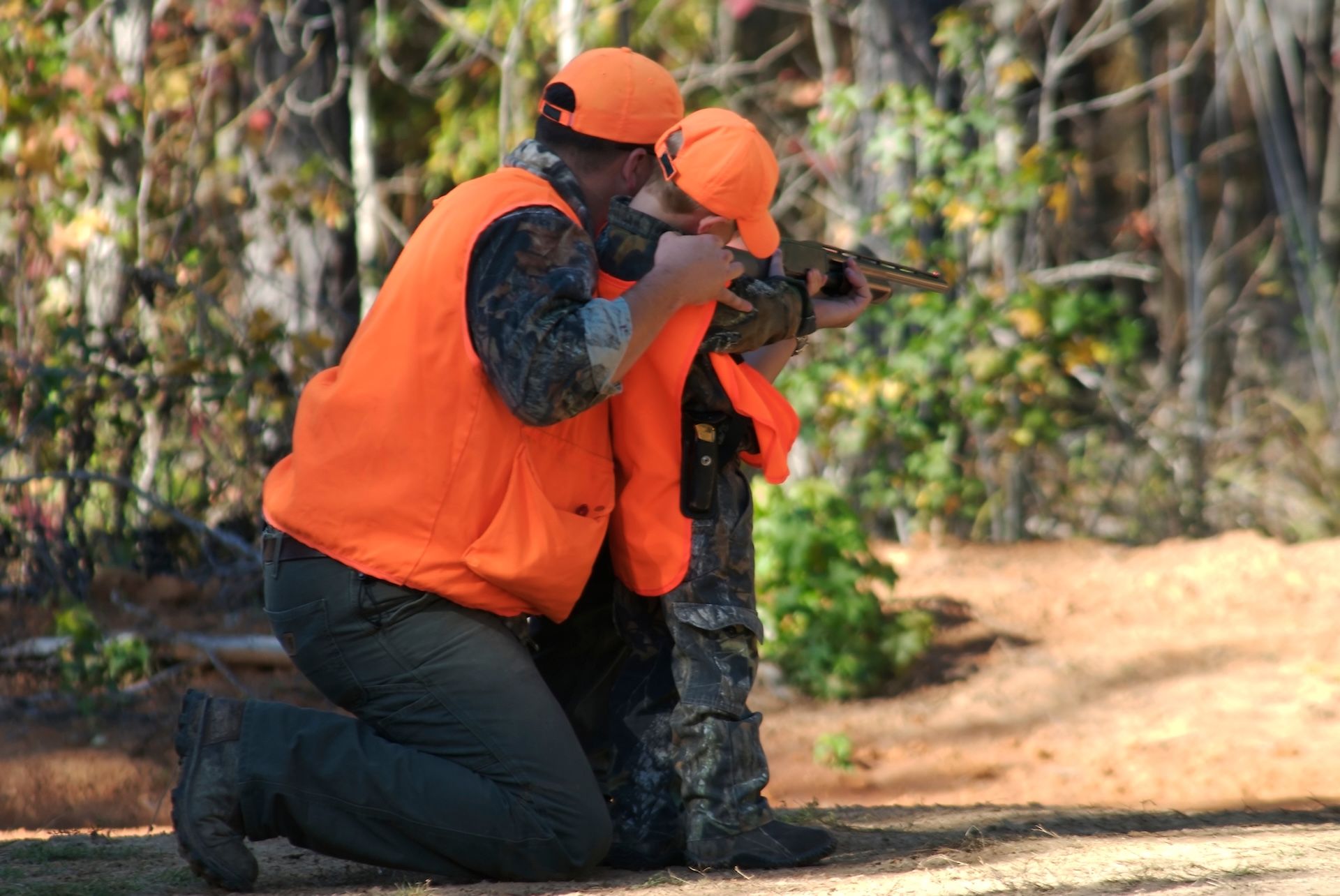 Man and boy in blaze orange aim rifle into woods. 