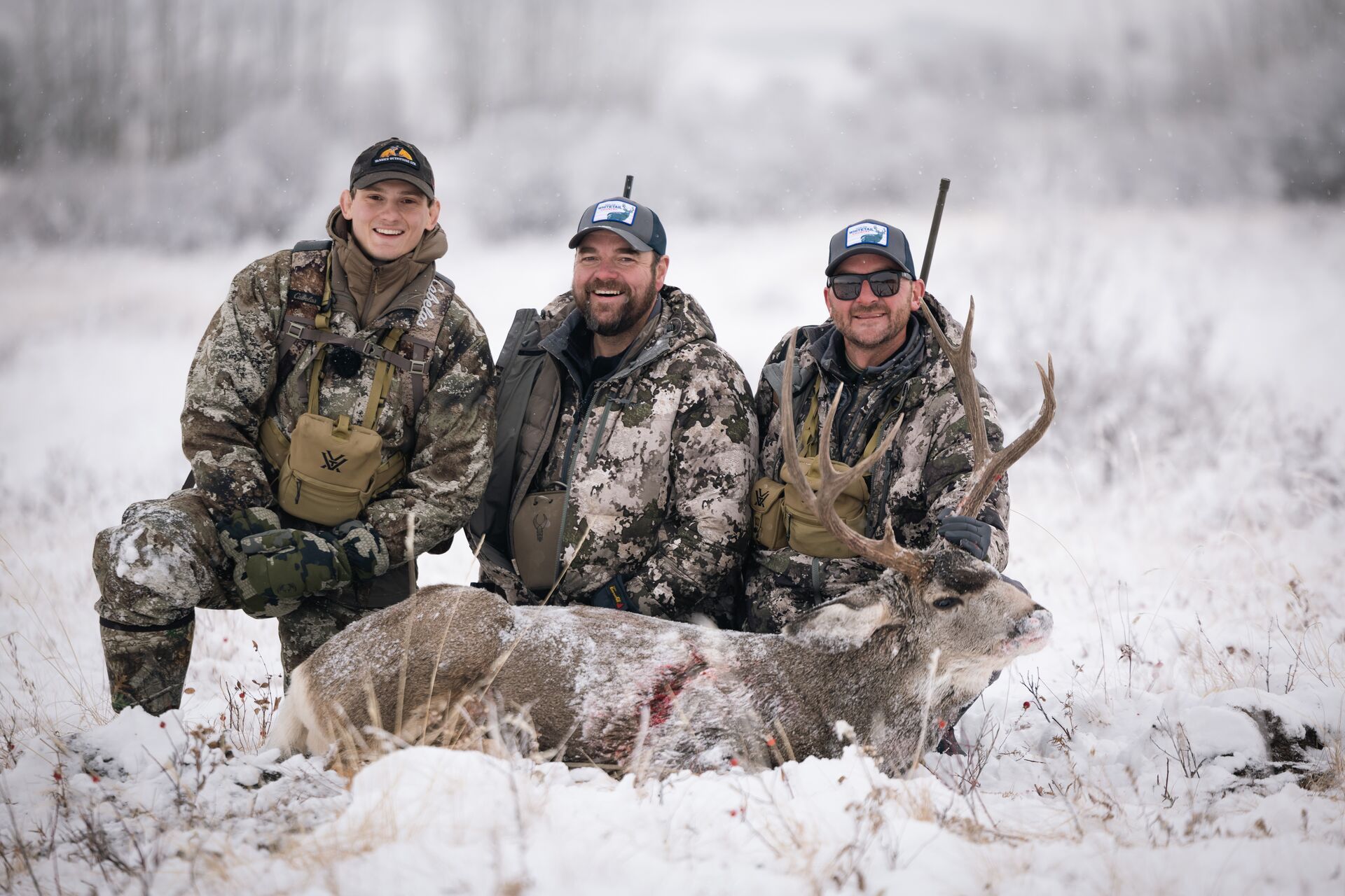 Three hunters in the snow with rifles and buck after hunt, best whitetail deer hunting rifle concept. 