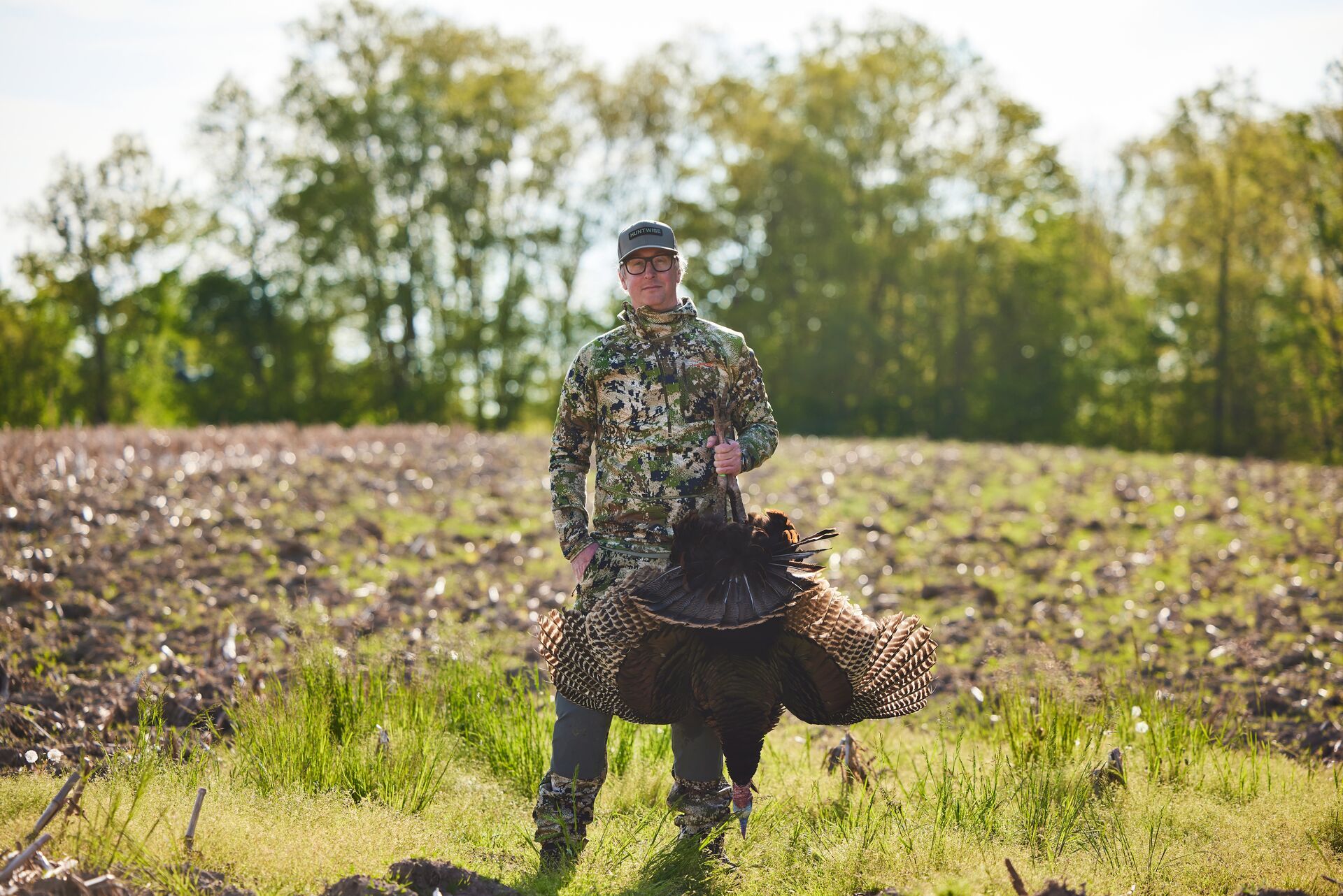 A smiling hunter in camo holds up a turkey after turkey hunting. 