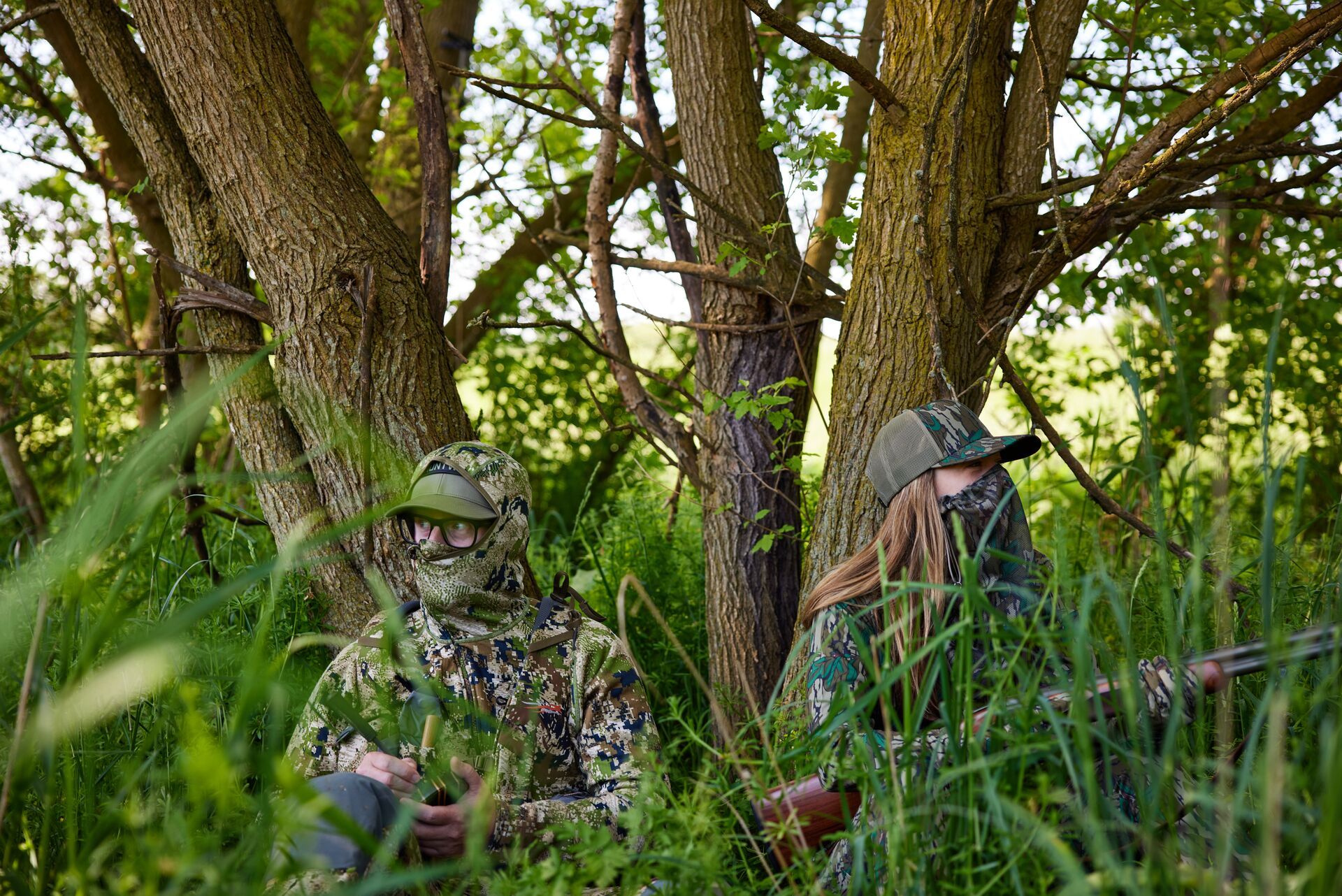 Two hunters in turkey camo blending into the trees and tall grass. 