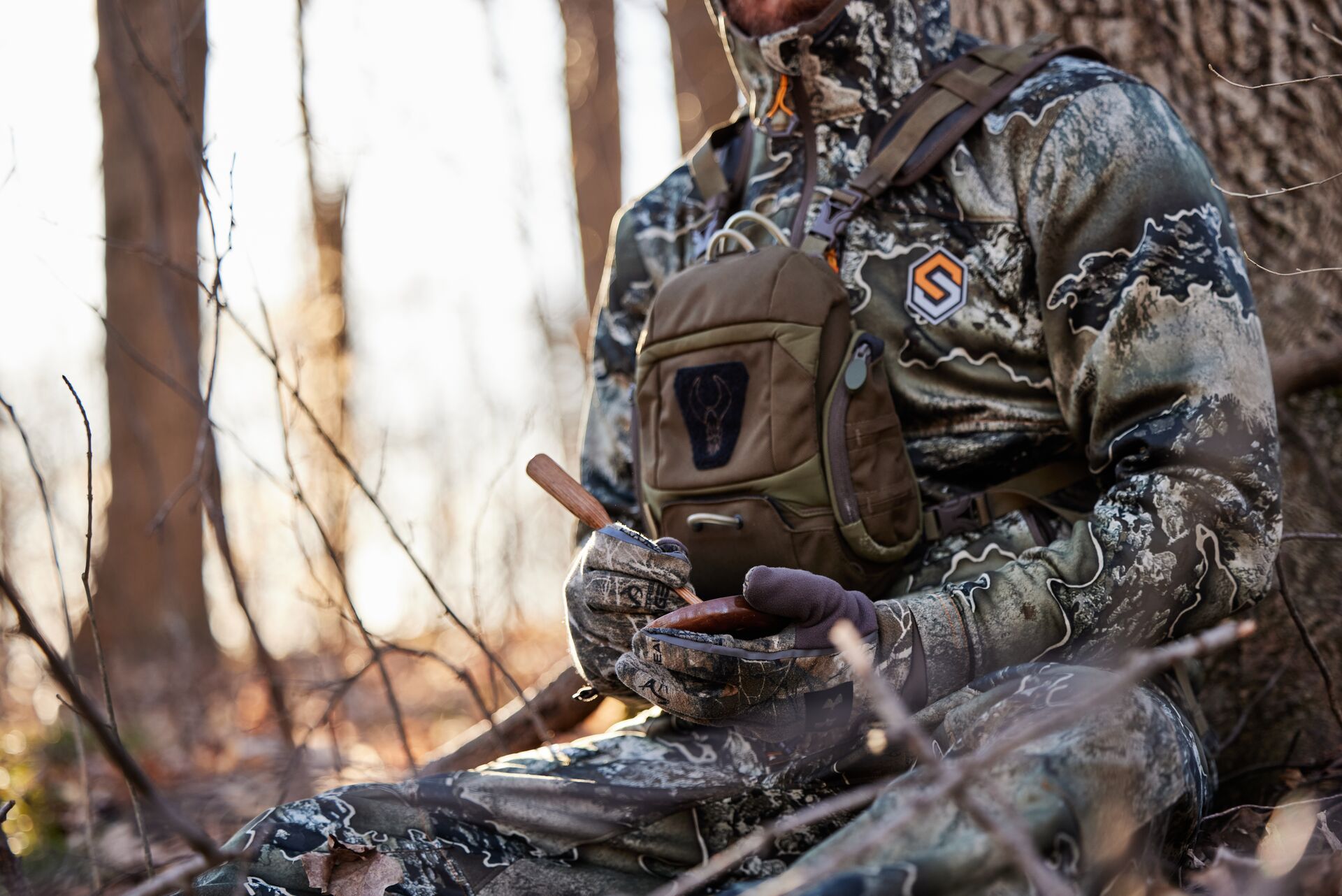 A hunter in camo sits against a tree using a turkey call, hunt turkey concept. 