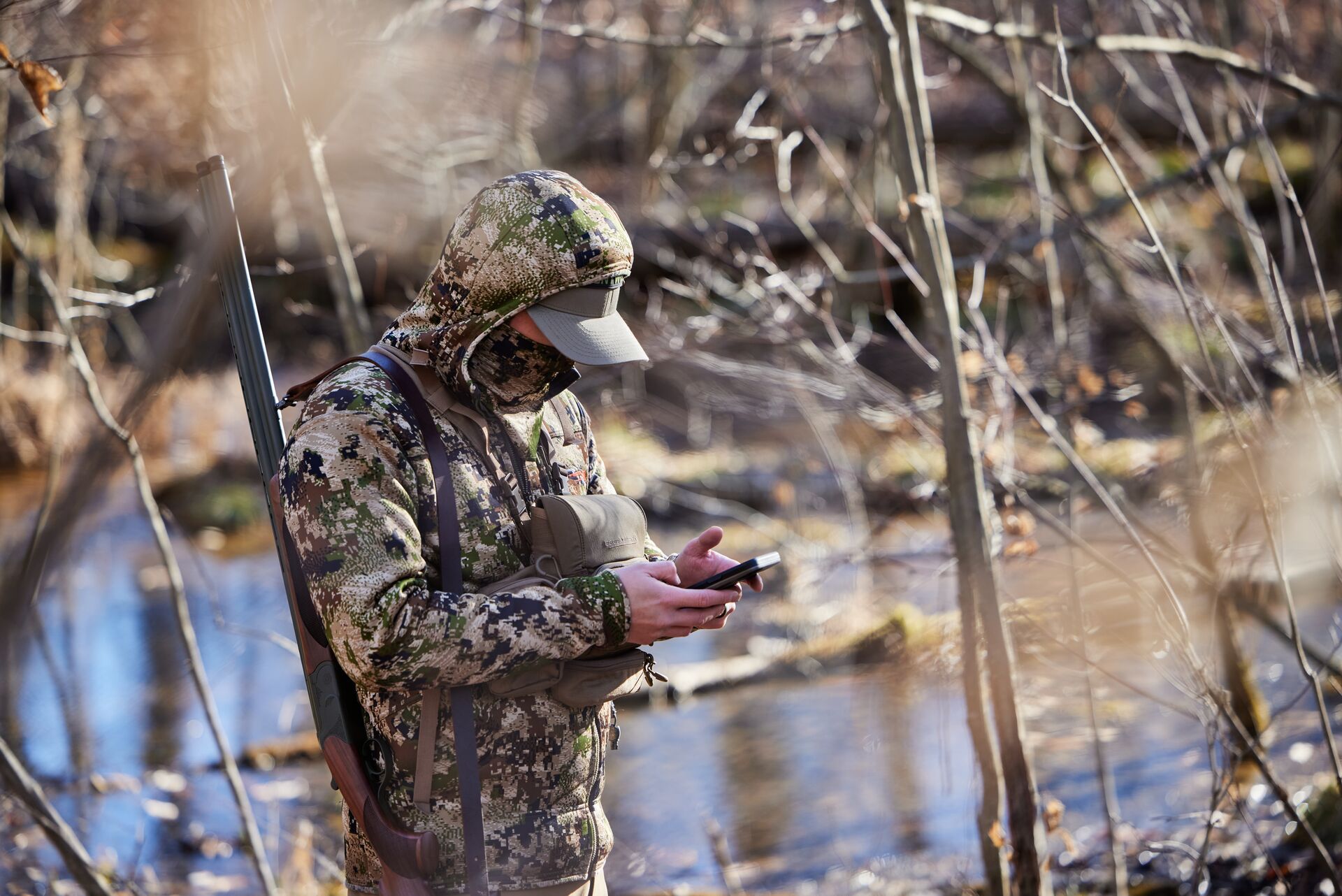 A hunter in camo with a shotgun looking at a phone, use HuntWise for Pennsylvania turkey hunting concept. 