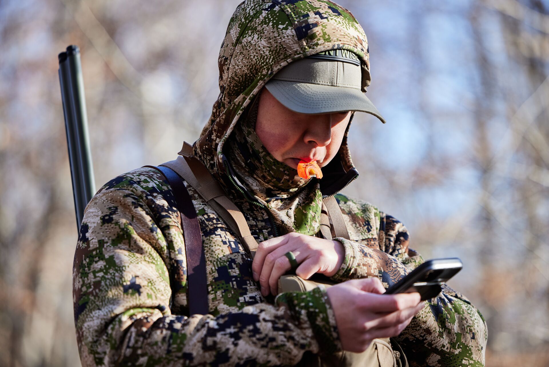 A hunter with turkey call in his mouth holding a shotgun and looking at a phone, use HuntWise to hunt Gould's turkey concept. 