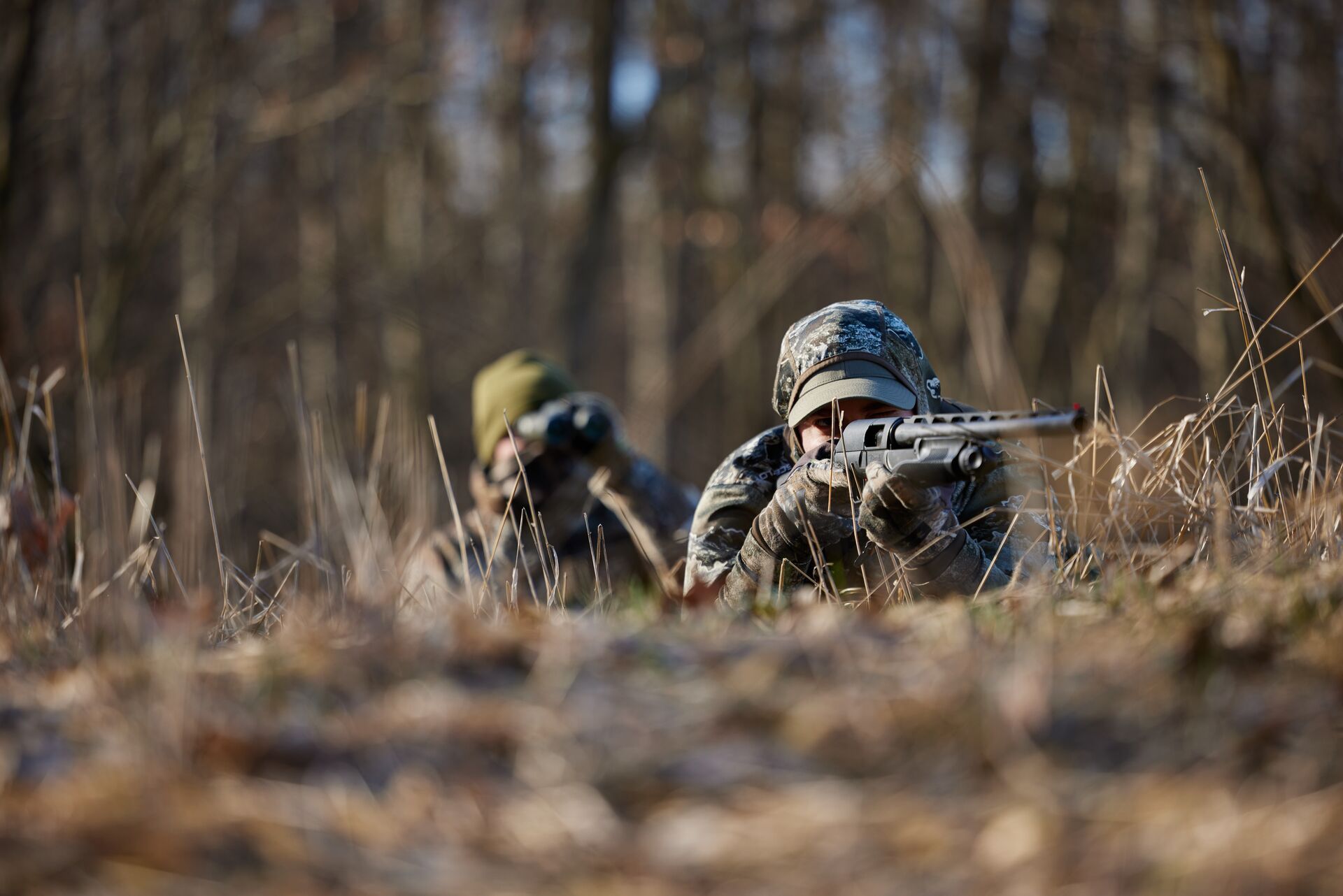 A hunter in camo on the ground aiming a shotgun with a second hunter spotting behind, hunting techniques concept. 