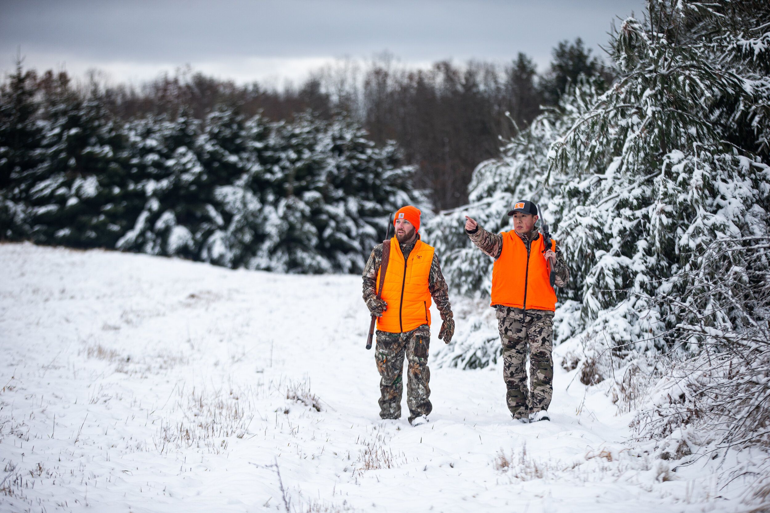 Two hunters in blaze orange and cold-weather gear walk through snow for late-season whitetail hunting.