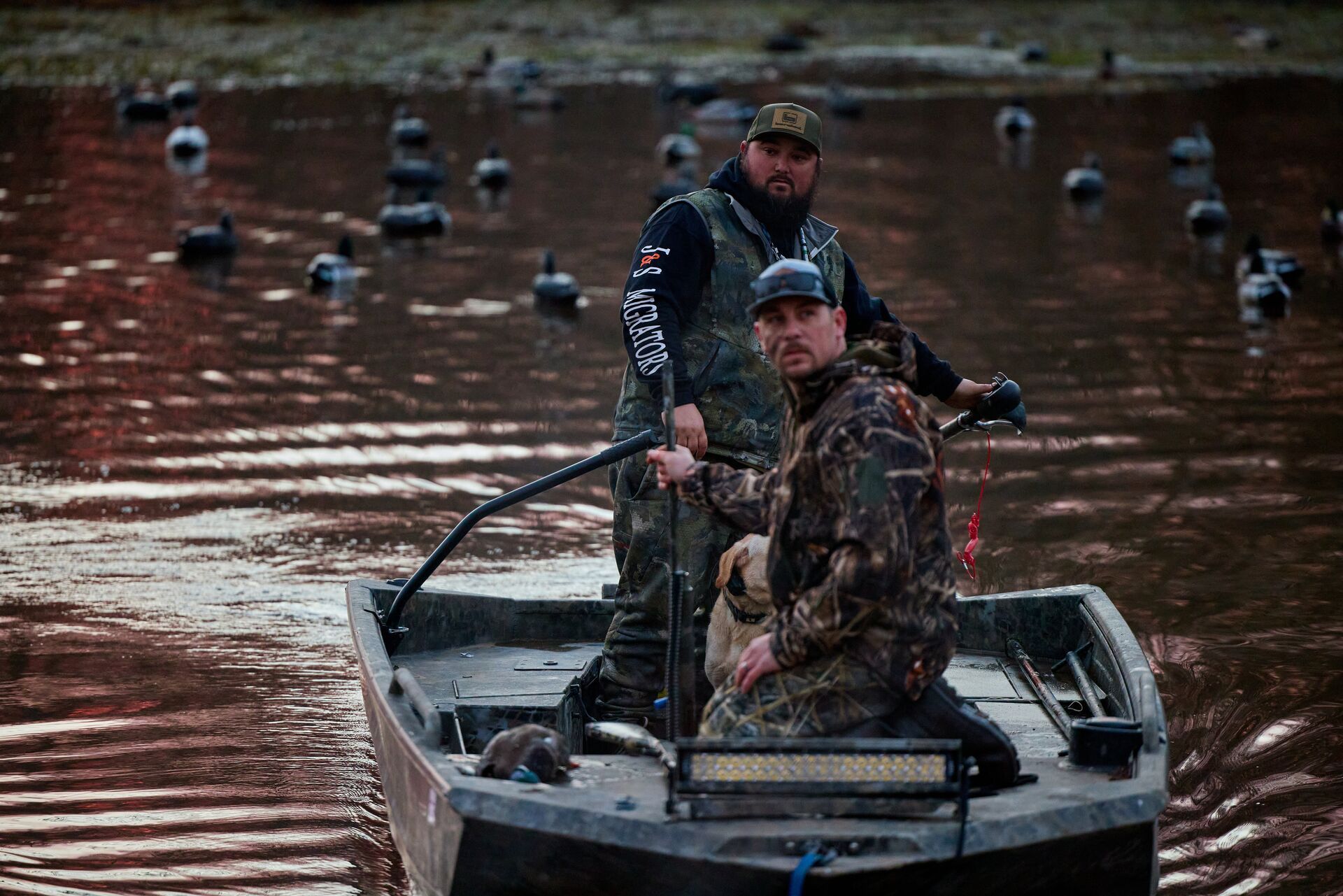 Two waterfowl hunters on a boat near decoys on the water. 